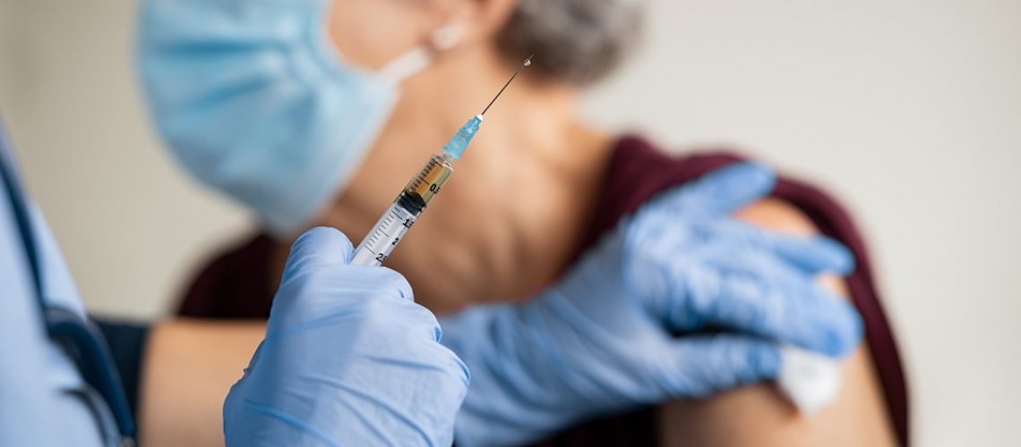 Close up of general practitioner hand holding vaccine injection while wearing face protective mask during covid-19 pandemic. Young woman nurse with surgical mask giving injection to senior woman at hospital. Close up of nurse holding syringe to vaccine old patient from covid19.